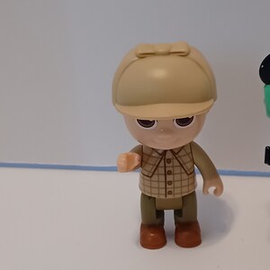 Puede incluir: Una peque&ntilde;a figura de pl&aacute;stico de una persona que lleva una camisa a cuadros marr&oacute;n y beige, pantalones marrones y un sombrero beige con un borde marr&oacute;n. La figura tiene una tez marr&oacute;n claro y ojos marrones.