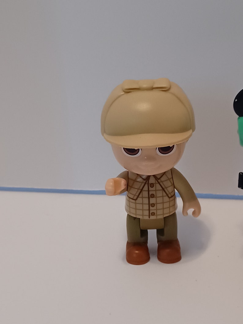 Puede incluir: Una peque&ntilde;a figura de pl&aacute;stico de una persona que lleva una camisa a cuadros marr&oacute;n y beige, pantalones marrones y un sombrero beige con un borde marr&oacute;n. La figura tiene una tez marr&oacute;n claro y ojos marrones.