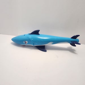 Può includere: Un giocattolo a forma di squalo blu chiaro con pinne e coda blu scuro. Lo squalo ha una striscia bianca lungo il fianco e un emblema rosso e bianco vicino all'occhio. Il giocattolo è di plastica.