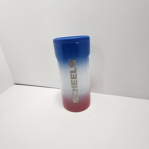 May include: A cylindrical tumbler with a blue lid and a gradient design. The top is blue, transitioning to white, and ending with a red base. The word "SCHEELS" is printed vertically in silver.
