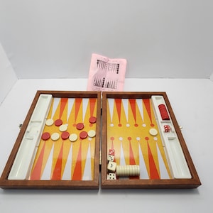 Peut inclure: Jeu de backgammon ouvert avec des pions rouges et blancs, des dés et un cube de doublement. Le plateau a un cadre en bois et une surface de jeu jaune et orange. Une feuille d'instructions rose est visible.