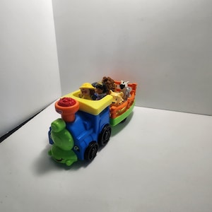 Peut inclure: Un train jouet coloré avec une locomotive bleue, un toit jaune et des accents verts. Le wagon transporte de petites figurines d'animaux et deux figurines de personnes. Le train a des roues noires et une cheminée orange.