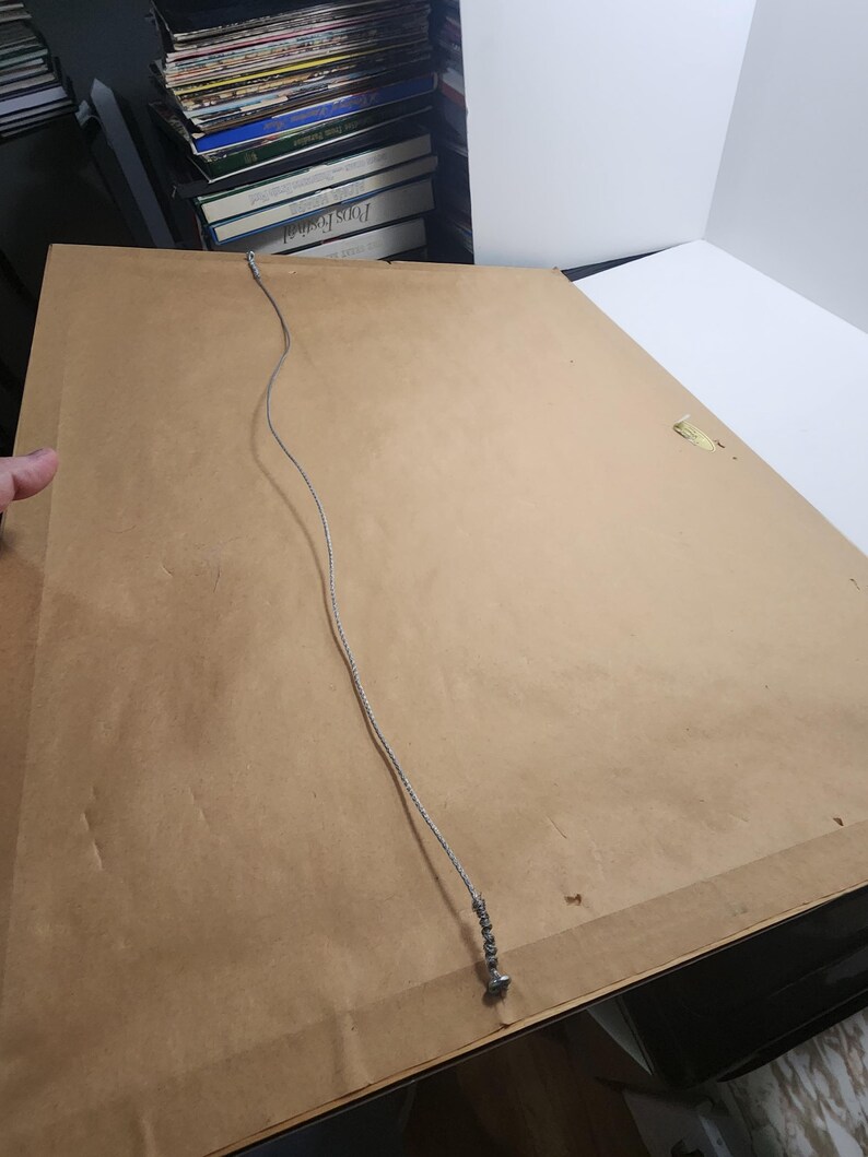 Peut inclure: Dos d'une &oelig;uvre encadr&eacute;e. Le dos est en carton brun clair. Un fil d'argent est attach&eacute; pour l'accrochage. L'&oelig;uvre est pos&eacute;e sur une surface blanche. Une main est visible sur le c&ocirc;t&eacute; gauche du cadre.