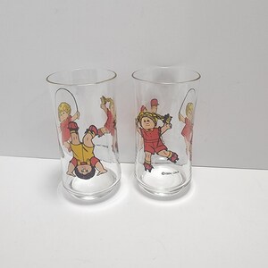 May include: Two clear glass drinking glasses with a cartoon image of a girl and boy jumping rope. The image includes the text "©1984 O.A.A."