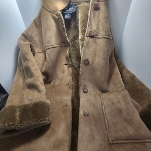 May include: A brown suede coat with a faux fur lining and trim. The coat has a button-up front, a notched collar, and two front pockets. The sleeves have a faux fur cuff. The coat is a medium size.
