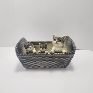 Puede incluir: Una cesta gris de cerámica con textura tejida, que contiene tres pequeñas figuras de gato detalladas. Los gatos tienen pelaje gris y blanco, ojos verdes y detalles rosas en las orejas. La cesta es rectangular y está sobre una superficie blanca.