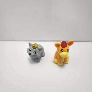 May include: Two small plastic animal figurines. One is a gray cat with a gold-colored crown. The other is an orange and yellow calf with a red gem on its head. Both are sitting on a white surface.