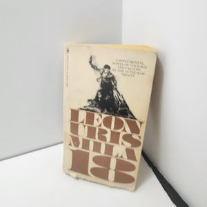 May include: A paperback book titled Mila 18 by Leon Uris with a cream-colored cover. The cover features a black silhouette illustration of a person holding a torch above a crowd, with the title and author name in large, bold brown lettering.