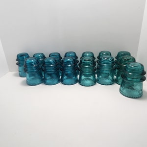 May include: A collection of 14 teal blue glass insulators. The insulators are all the same size and shape, and they have a smooth, rounded top. They are arranged in a row on a white surface.