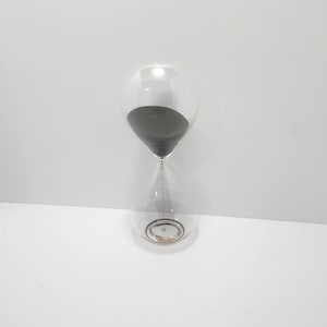 May include: A clear glass hourglass with black sand inside. The hourglass has a bulbous top and bottom, connected by a narrow neck. The sand is partially through the hourglass, indicating the passage of time. The hourglass is set against a white background.