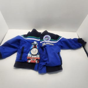Puede incluir: Una chaqueta azul y azul marino con cremallera que presenta un gráfico de Thomas el tren en la parte delantera. La chaqueta incluye un parche circular con el texto 'Thomas the Train Sodor Engine Co. No. 1' y el nombre 'Thomas' bordado.