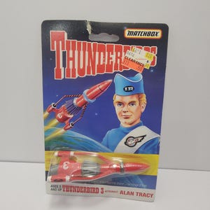 Puede incluir: Juguete Matchbox Thunderbird 3 en una tarjeta azul y negra. La tarjeta presenta una imagen de dibujos animados de un hombre con uniforme azul y un cohete rojo. El juguete es rojo y blanco. El texto en la tarjeta incluye "Thunderbirds" y "Alan Tracy".