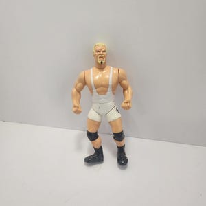 May include: A vintage action figure of a muscular wrestler. The figure has blonde hair, a goatee, and is wearing a white singlet with suspenders, black knee pads, and black boots. The figure is posed with arms at its sides.