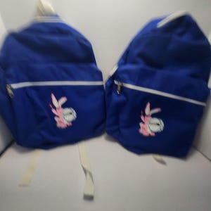 May include: Two blue backpacks with white trim and straps. Each backpack features a pink and white cartoon bunny graphic on the front. The backpacks have a zippered closure and a top handle.