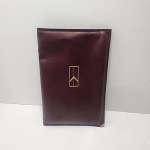 May include: A burgundy leather portfolio with a gold emblem in the upper center. The portfolio is rectangular and appears to be closed. The emblem is a stylized geometric design.