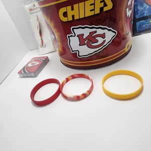 May include: A collection of Kansas City Chiefs merchandise, including a red and gold bucket, playing cards, and three rubber wristbands. One wristband is red, one is yellow, and one is a mix of red, yellow, and orange.