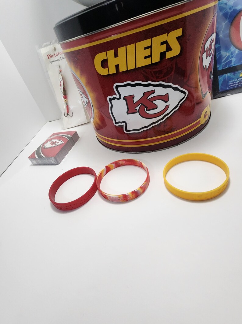 May include: A collection of Kansas City Chiefs merchandise, including a red and gold bucket, playing cards, and three rubber wristbands. One wristband is red, one is yellow, and one is a mix of red, yellow, and orange.