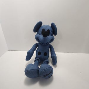 Puede incluir: Un peluche de Mickey Mouse de estilo denim. El peluche presenta una tela de mezclilla azul con detalles negros para los ojos, la nariz y los botones. El logotipo de Disney es visible en el pie. El juguete est&aacute; sentado.