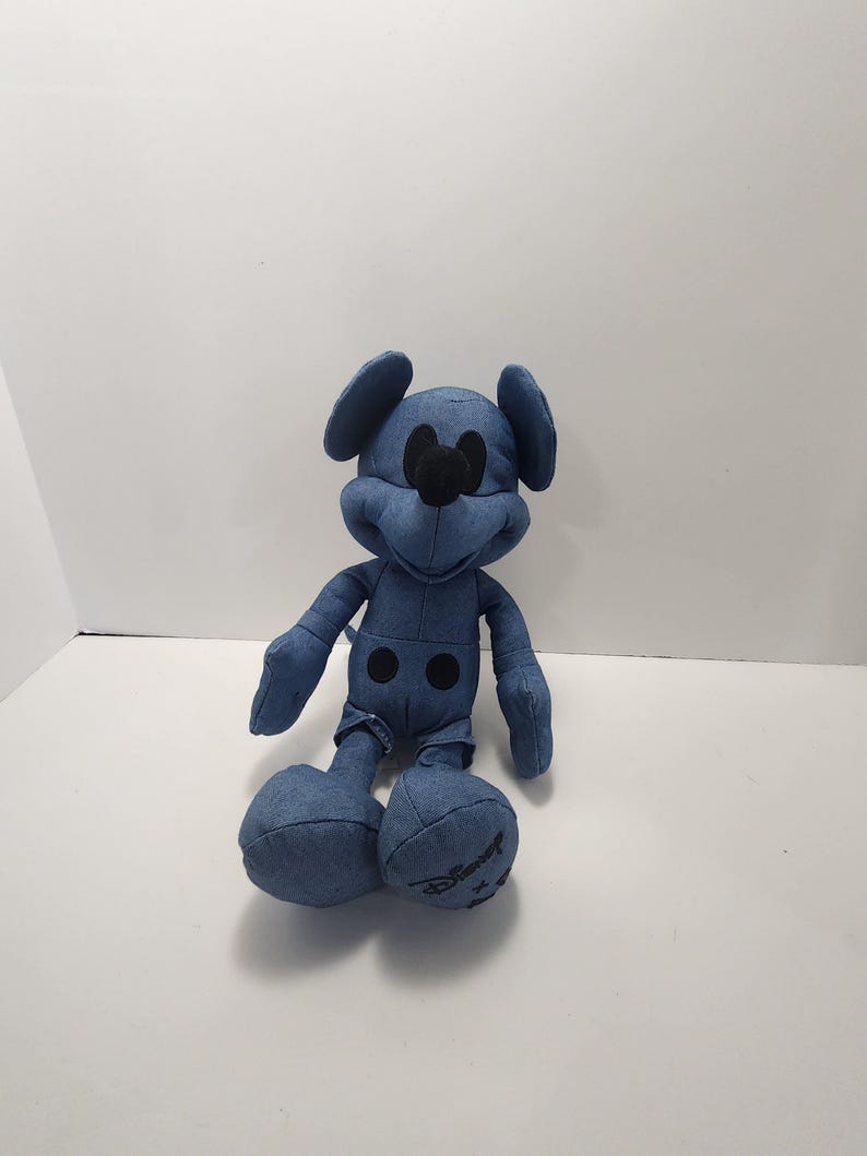 Puede incluir: Un peluche de Mickey Mouse de estilo denim. El peluche presenta una tela de mezclilla azul con detalles negros para los ojos, la nariz y los botones. El logotipo de Disney es visible en el pie. El juguete est&aacute; sentado.