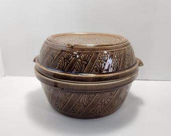 Può includere: Una casseruola in ceramica marrone con coperchio. La casseruola ha un design decorativo di foglie e volute stilizzate.
