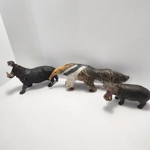 May include: Three plastic animal figurines: a brown and black anteater, and two black hippos with open mouths. The anteater is standing on all fours, while the hippos are lying down.
