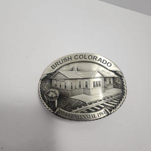 May include: Silver-toned metal belt buckle featuring a detailed relief of a building with the text "BRUSH COLORADO" and "1884 CENTENNIAL 1984". The buckle has a textured border and depicts railroad tracks.