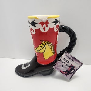 Puede incluir: Taza de cerámica con forma de bota de vaquero, con asa negra y caña roja con cabeza de caballo amarilla. La bota tiene puntera negra y diseño de estrellas blancas y negras en la parte superior. Una etiqueta dice "Vintage Cowboy Boot Mugs".