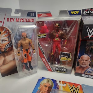 Op de afbeelding: Vier ongeopende actieve figuren van professionele worstelaars. De figuren zijn Rey Mysterio, Stevie Ray, Goldberg en Ric Flair. De figuren zijn verpakt in hun originele dozen. De dozen zijn rood, zwart en wit met het WWE-logo.
