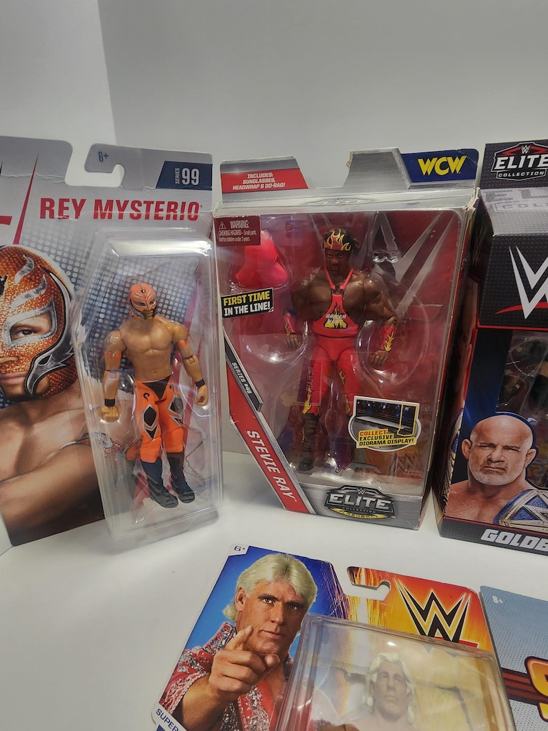 Op de afbeelding: Vier ongeopende actieve figuren van professionele worstelaars. De figuren zijn Rey Mysterio, Stevie Ray, Goldberg en Ric Flair. De figuren zijn verpakt in hun originele dozen. De dozen zijn rood, zwart en wit met het WWE-logo.