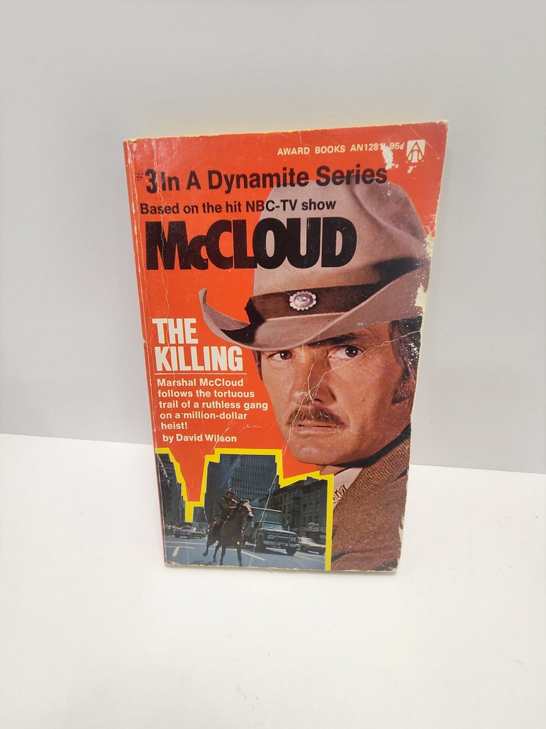 May include: A red and white paperback book cover with the title "The Killing" and the text "Marshal McCloud follows the tortuous trail of a ruthless gang on a million-dollar heist!"  The cover features a photo of a man wearing a brown cowboy hat and a brown jacket. The book is part of the "3 In A Dynamite Series" based on the NBC-TV show "McCloud".