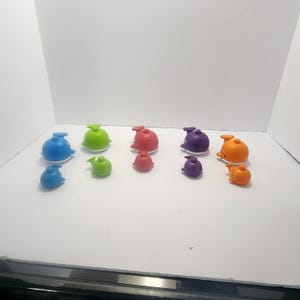 Puede incluir: Una colección de juguetes de baño con forma de ballena de colores en varios tamaños. Los juguetes son en tonos de azul, verde, rojo, morado y naranja. Algunos tienen números. Los juguetes están colocados sobre una superficie blanca.