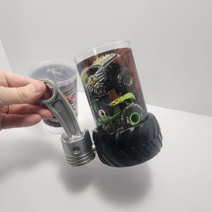 May include: A clear glass tumbler featuring a monster truck design, with a black tire base. A silver piston is next to the tumbler. The tumbler has a colorful graphic of a monster truck with green and yellow accents.