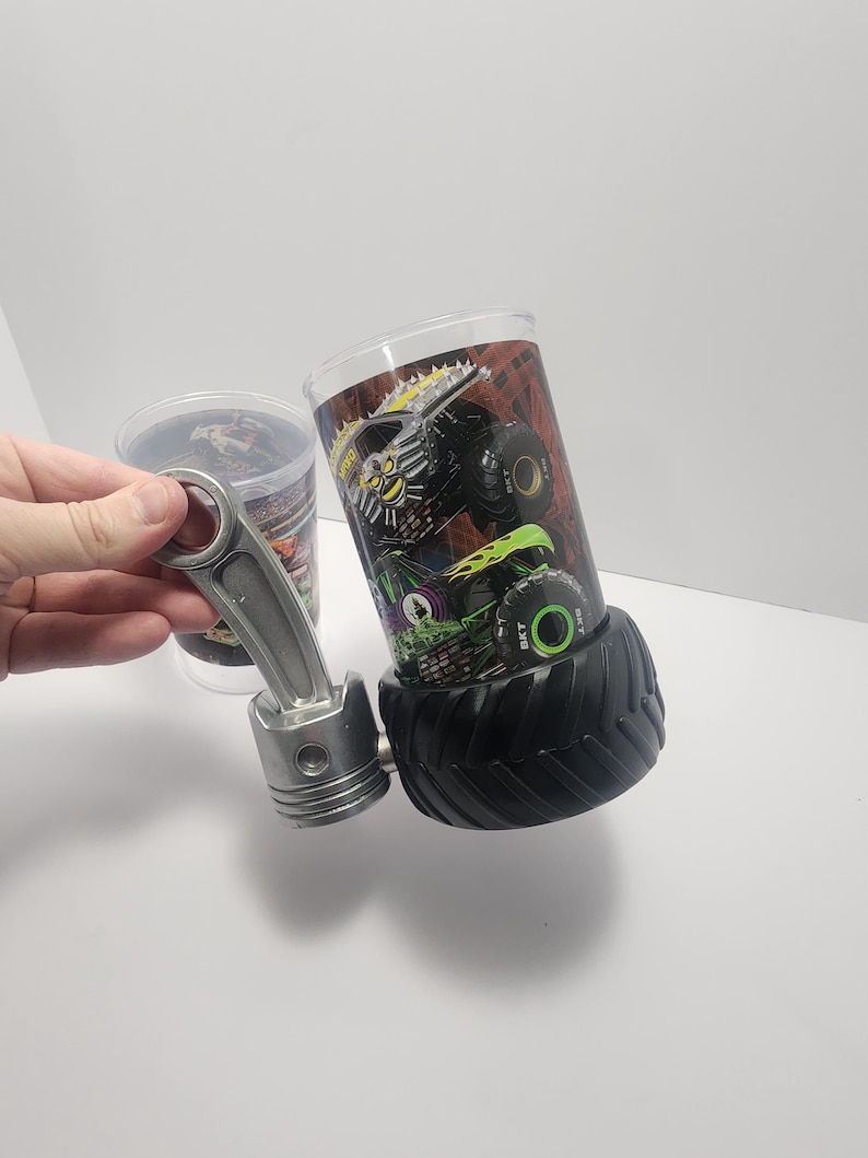 May include: A clear glass tumbler featuring a monster truck design, with a black tire base. A silver piston is next to the tumbler. The tumbler has a colorful graphic of a monster truck with green and yellow accents.