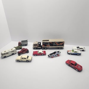 May include: A collection of toy cars and trucks, including a semi-truck with a camouflage trailer, a red convertible, a white camper, and several other miniature vehicles. The toys are in various colors and styles, arranged on a white surface.