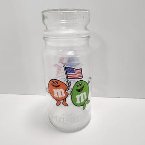 May include: A clear glass jar with a lid featuring an American flag, an orange M&M character, and a green M&M character holding the flag. The jar has the words "M&M's Candies" printed on the lid.
