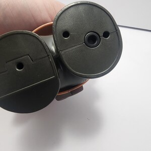 May include: A dark brown, plastic object with two circular, hinged components. Each component has a small hole and a raised edge. The object appears to be a part of a larger mechanism or device.