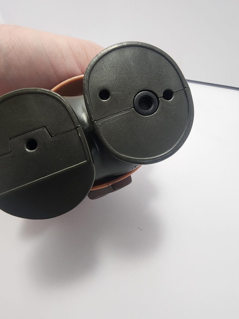 May include: A dark brown, plastic object with two circular, hinged components. Each component has a small hole and a raised edge. The object appears to be a part of a larger mechanism or device.