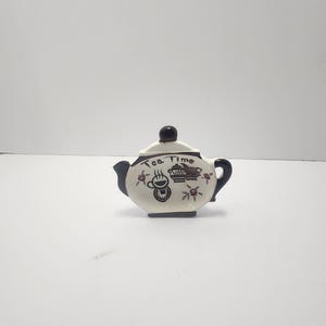 Puede incluir: Pequeña tetera de cerámica blanca con detalles negros. La tapa y el asa son negras. Decorada con diseños florales y de café negros y rojos, con la inscripción "Tea time" en la tapa.