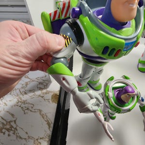 May include: A green and white Buzz Lightyear action figure with articulated arms and legs. The figure has a purple chest piece and a clear plastic visor. The figure is holding a smaller Buzz Lightyear action figure in its right hand.