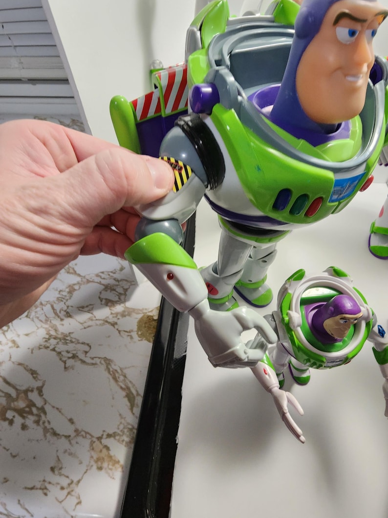 May include: A green and white Buzz Lightyear action figure with articulated arms and legs. The figure has a purple chest piece and a clear plastic visor. The figure is holding a smaller Buzz Lightyear action figure in its right hand.