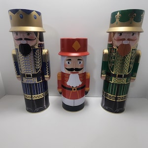 May include: Three vintage-style nutcracker tin containers. The nutcrackers are dressed in blue, red, and green uniforms with gold accents. The containers are ideal for storing small items or as decorative accents.