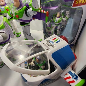 May include: A white and blue toy spaceship with a green and purple wing. The spaceship has a clear cockpit with a small toy Buzz Lightyear figure inside. The spaceship is part of a Toy Story themed toy set.
