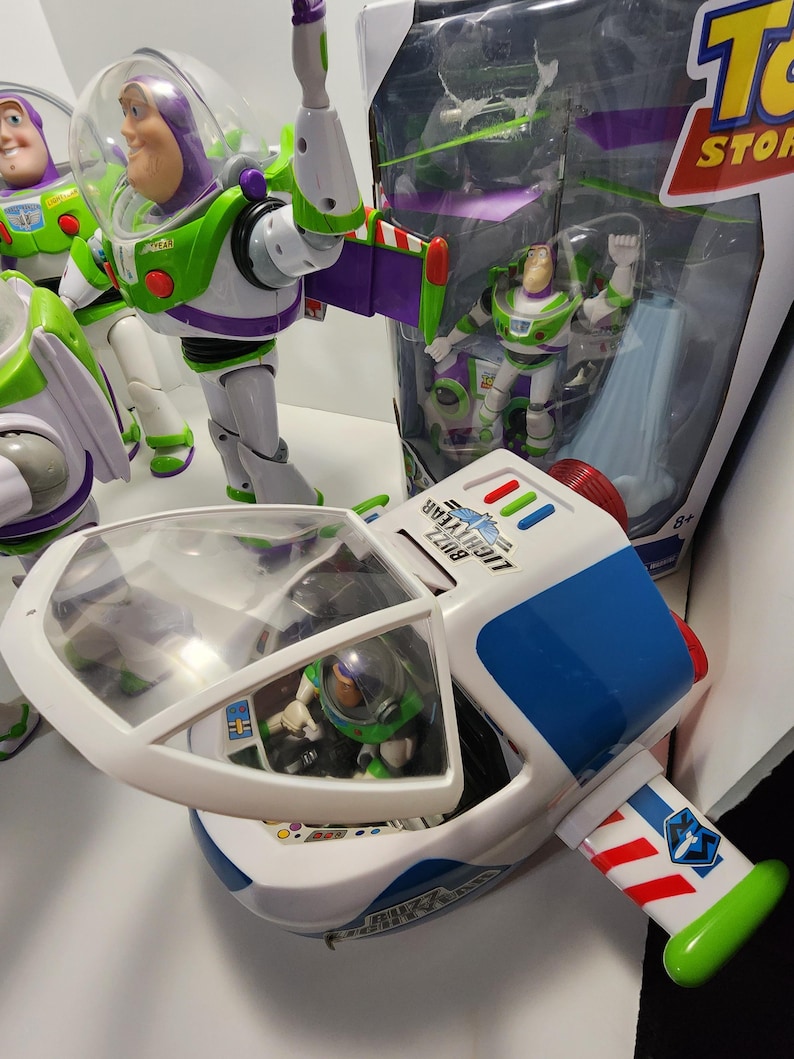 May include: A white and blue toy spaceship with a green and purple wing. The spaceship has a clear cockpit with a small toy Buzz Lightyear figure inside. The spaceship is part of a Toy Story themed toy set.