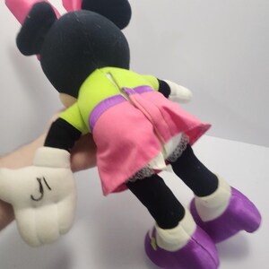 May include: A plush Minnie Mouse doll, featuring a black head with pink bow, black ears, and a lime green top with a pink skirt. The doll has white gloves with black markings and purple shoes. The doll is a classic Disney character.