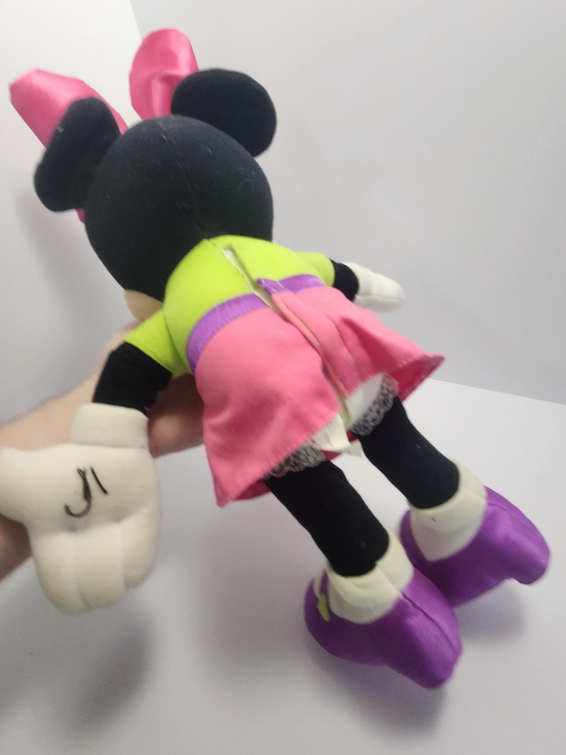 May include: A plush Minnie Mouse doll, featuring a black head with pink bow, black ears, and a lime green top with a pink skirt. The doll has white gloves with black markings and purple shoes. The doll is a classic Disney character.