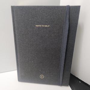 May include: A dark gray journal with the gold text "NOTE TO SELF" on the cover. The journal has a matching elastic band closure and a small circular emblem. The journal is a notebook for writing and note-taking.