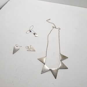 May include: A silver-toned jewellery set consisting of a necklace and two pairs of earrings. The necklace features five triangular metal pendants on a thin chain. One pair of earrings has small triangular drops, while the other pair features thin wire hooks with small, flat, leaf-shaped metal accents and tiny blue beads.