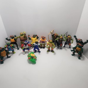 Op de afbeelding: Een verzameling Teenage Mutant Ninja Turtles-actionfiguren, waaronder Leonardo, Donatello, Raphael en Michelangelo. De figuren staan in verschillende poses en zijn gemaakt van plastic. Sommige figuren dragen verschillende outfits, waaronder een honkbaluniform en een clownkostuum.
