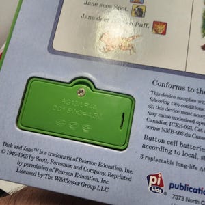 Puede incluir: Primer plano de un compartimento de bater&iacute;a verde en un libro infantil. El compartimento tiene el texto "AG13/LR44 DC1.5Vx3=4.5V". El texto del libro incluye "Dick and Jane" y "Run, run, run."