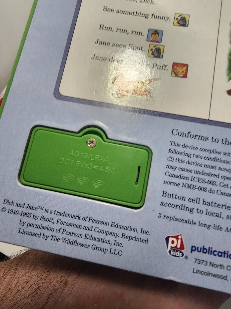 Puede incluir: Primer plano de un compartimento de bater&iacute;a verde en un libro infantil. El compartimento tiene el texto "AG13/LR44 DC1.5Vx3=4.5V". El texto del libro incluye "Dick and Jane" y "Run, run, run."